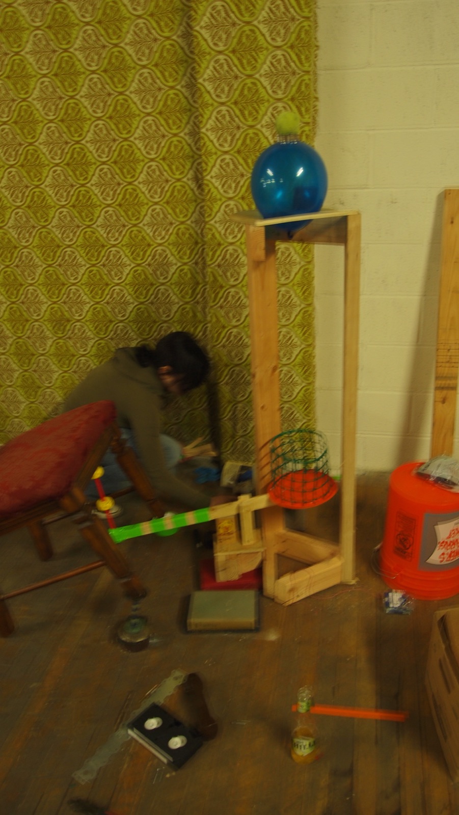 A tall wooden stand holding a blue ball, with a basket and wires at its base