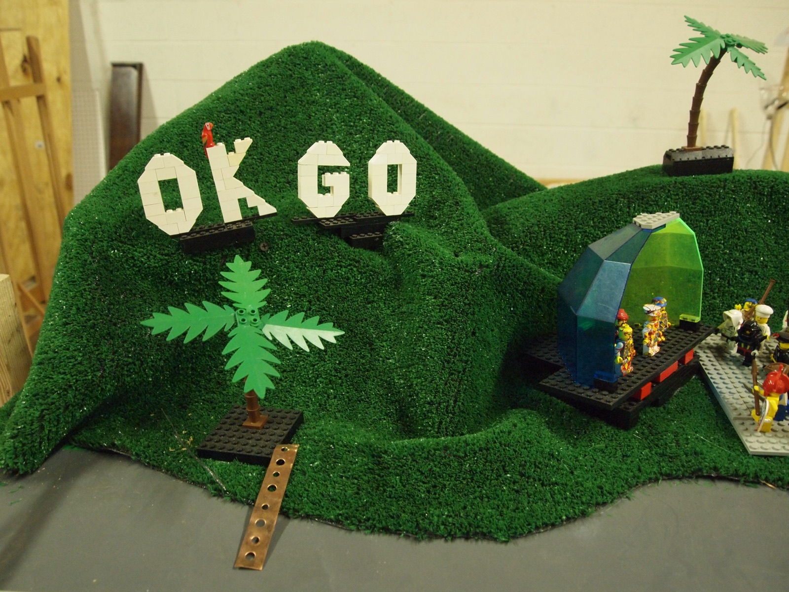 A small green hill model with the letters OK GO, a palm tree and toy figures