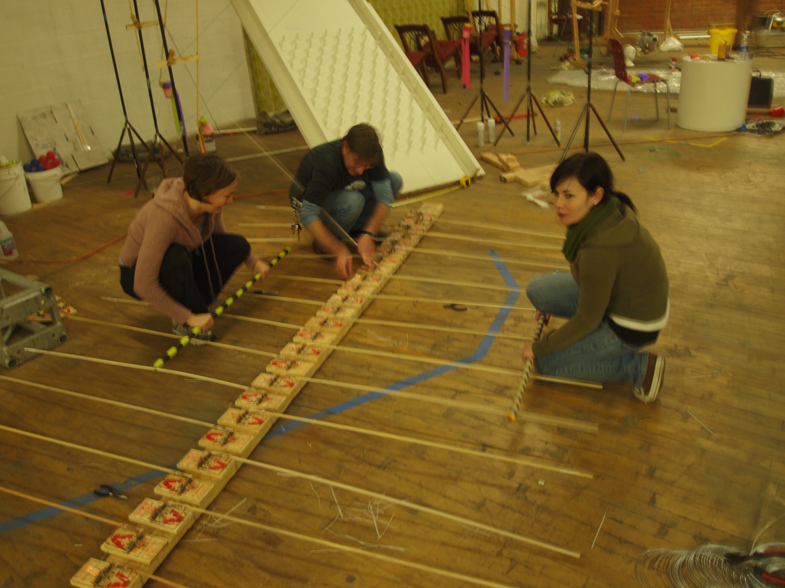 Three people kneeling on a wood floor setting up a line of mousetraps with wires